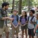 Monitor con mochila explica una actividad a cuatro niños en un campamento entre árboles