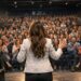 Un auditorio en el que una mujer da una conferencia motivacional, la imagen debe estar desde la perspectiva del escenario, con la mujer de espaldas en primer plano y la audiencia de fondo.